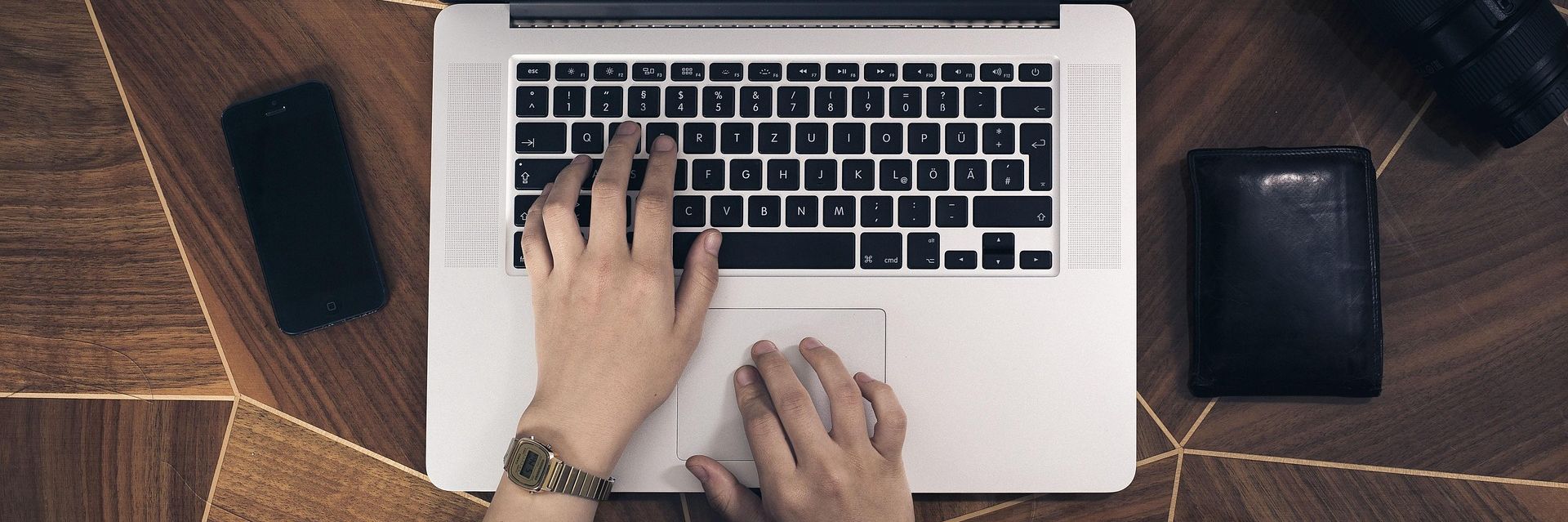 Ansicht von oben auf einen Laptop. Man sieht Hände. Links ein Handy. Rechts ein Portmonee