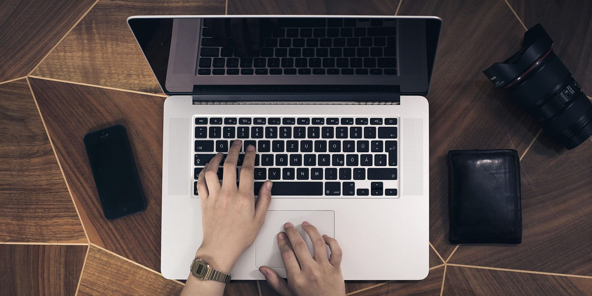 Ansicht von oben auf einen Laptop. Man sieht Hände. Links ein Handy. Rechts ein Portmonee
