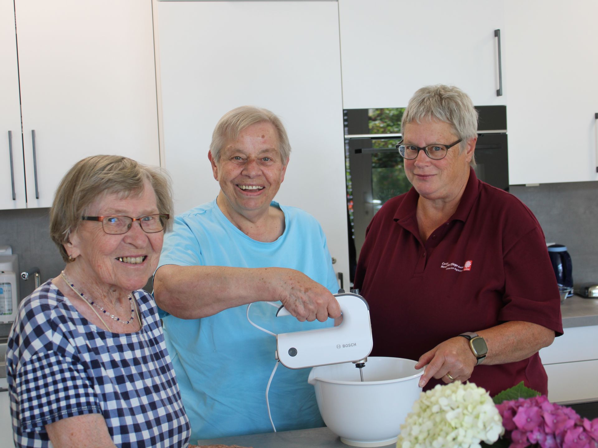 3 Mitarbeitende stehen in der Küche. Die in der Mitte hält den Mixer.