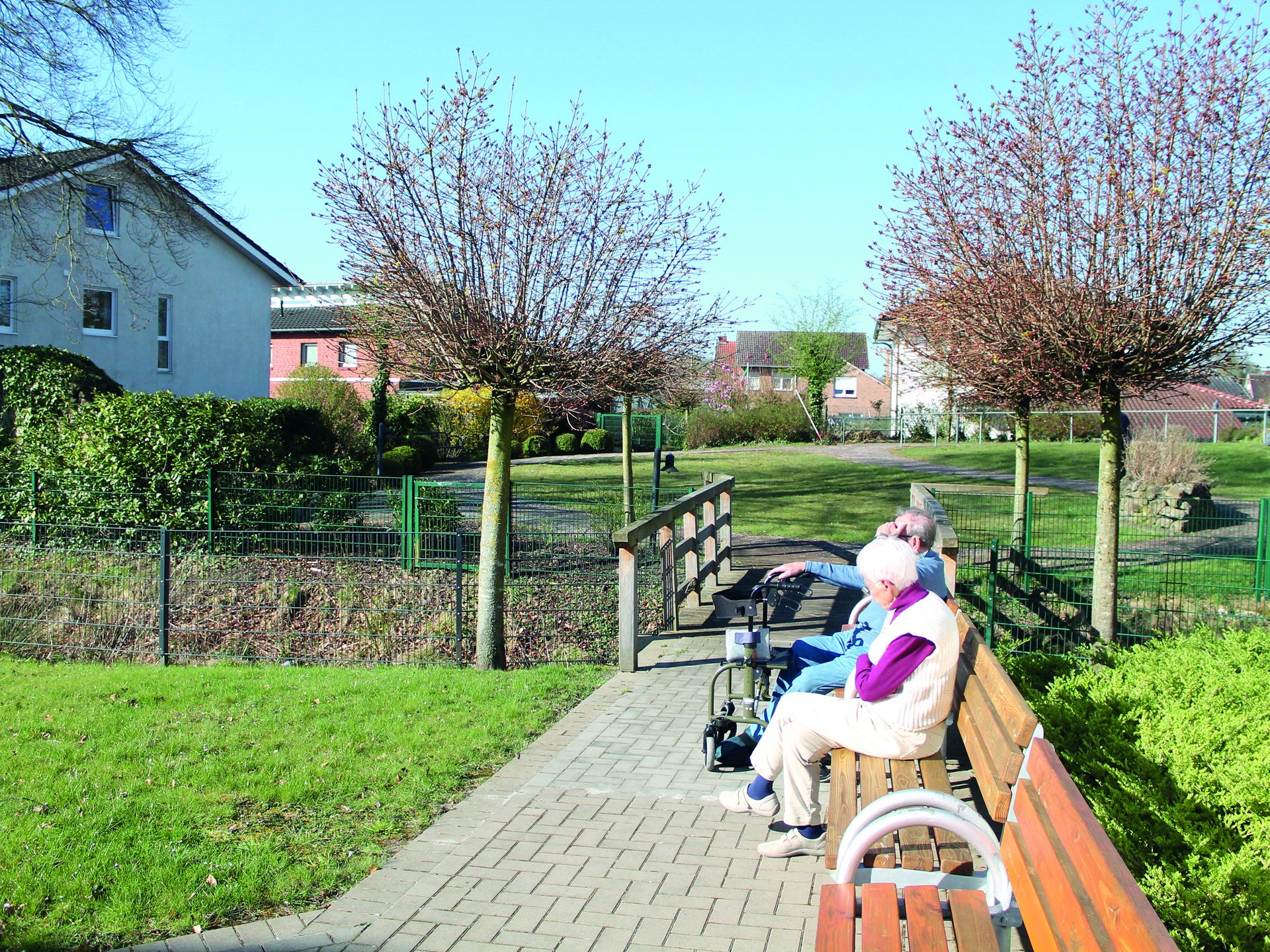 Bild im Garten. Rechts sitzen Personen auf Bänken. Im Hintergrund Büsche und Bäume.