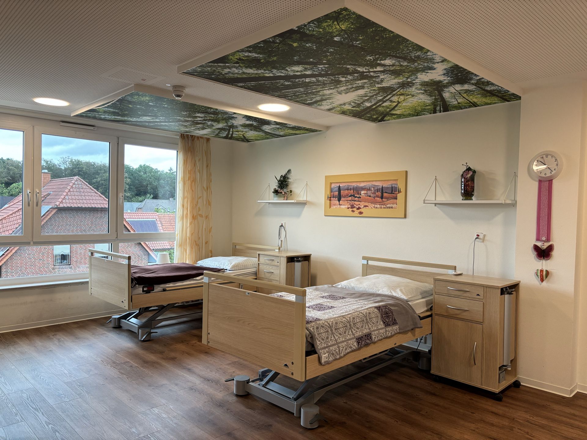 Zwei Pflegebetten mit Nachschrank stehen in einem Raum am Fenster. An der Decke hängt ein Bild und Deko an der Wand.