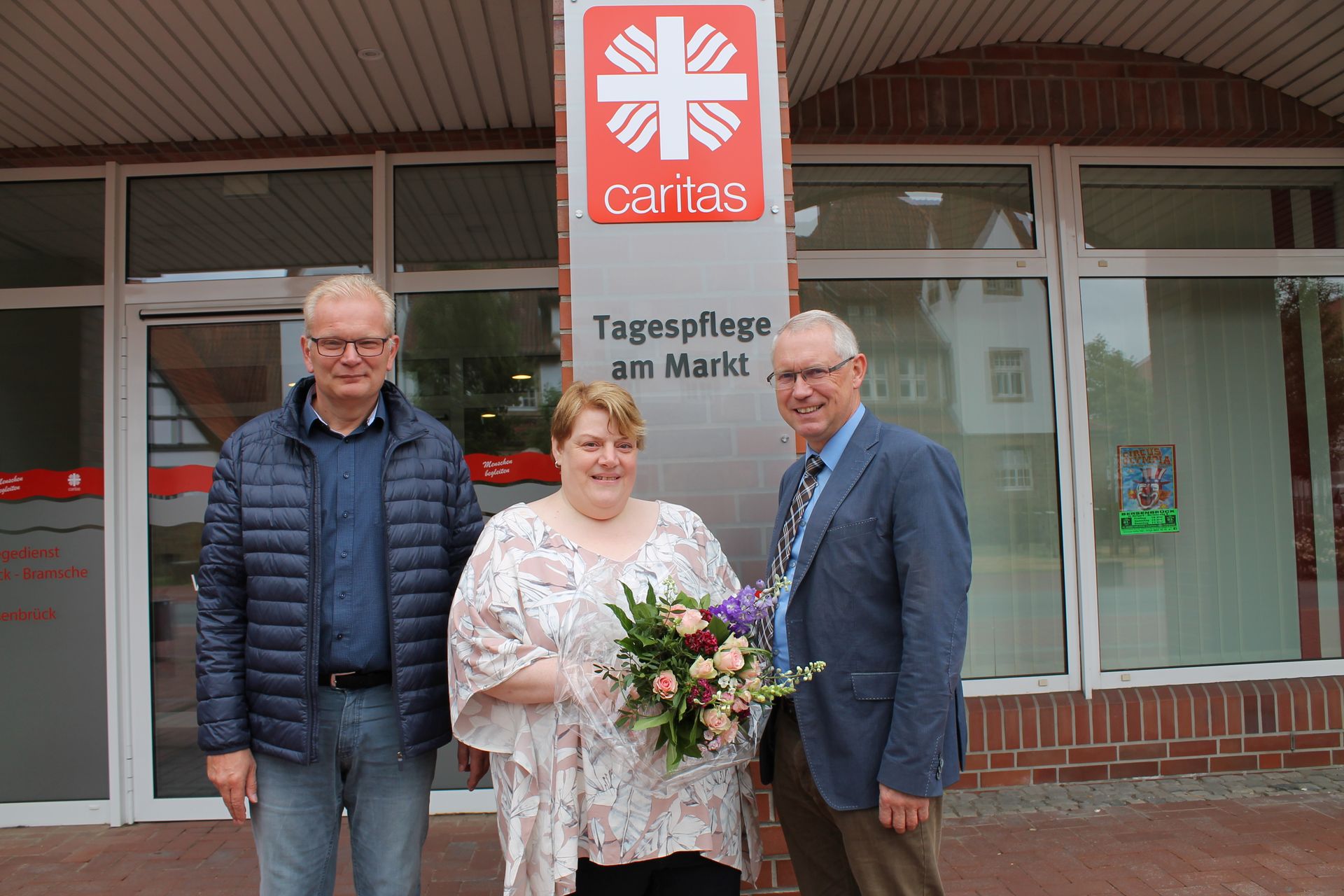 Drei Personen stehen vor einer Tagespflege. Die Frau in der Mitte trägt einen Blumenstrauß.